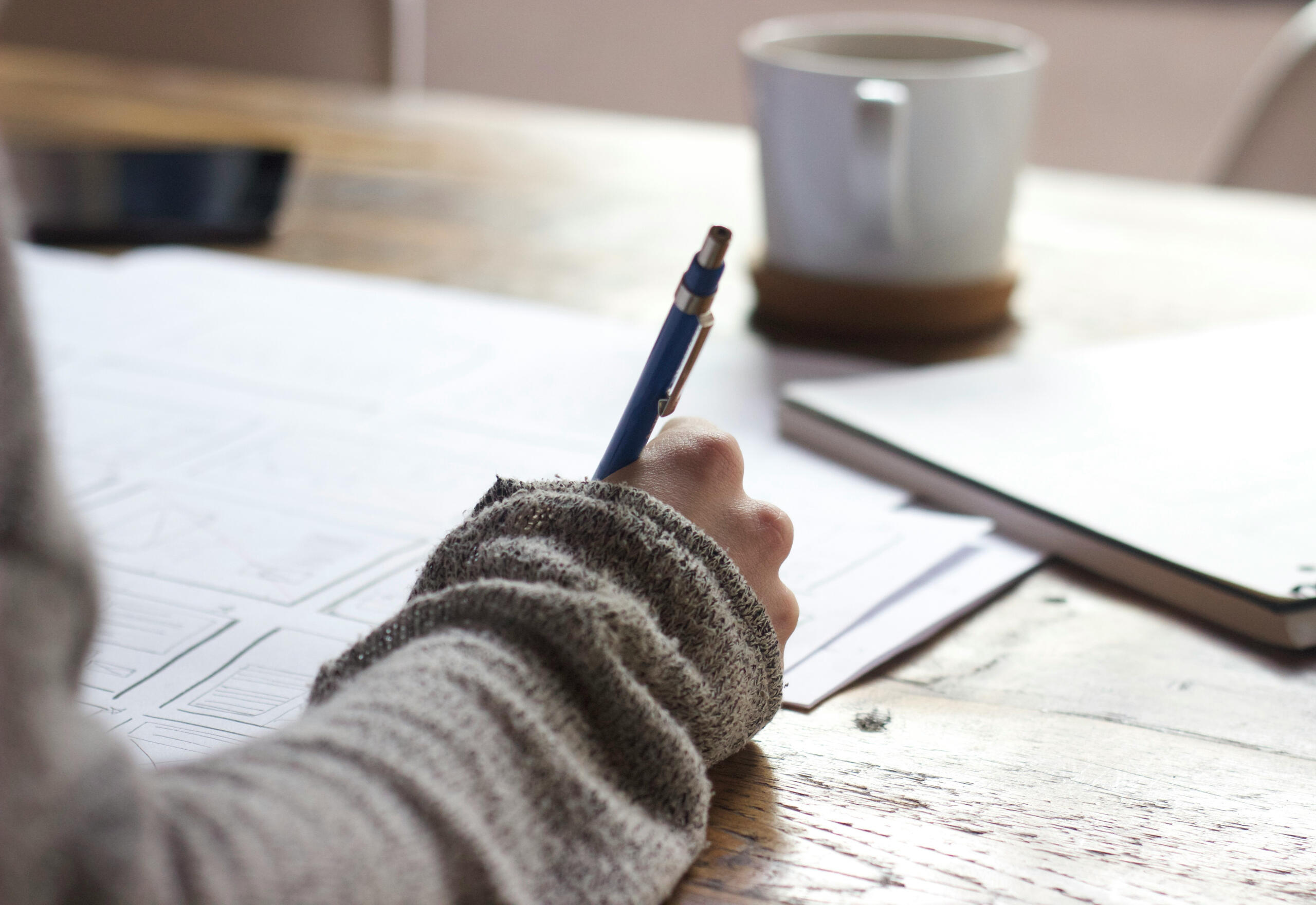 a closeup of someone's hand writing. they look like they are wearing a comfy sweater. there's a coffee nearby